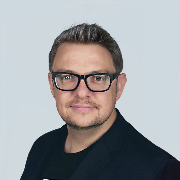 Man looking at camera, wearing a blue jacket and black t-shirt, wearing glasses