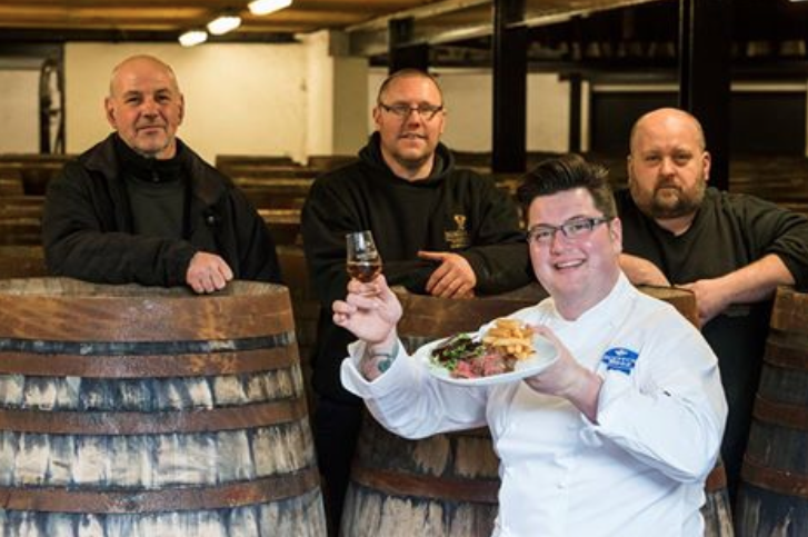 Three men standing over whisky barrels and one man in a chef outfit holding a glass of whisky and a plate of Scottish meat.