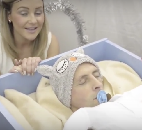 Man in crib with dummy in mouth, sleeping and woman with angel tinsel wings behind him