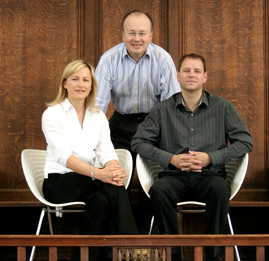 Group image of three people facing camera, two sat and one in between standing, two men and one woman