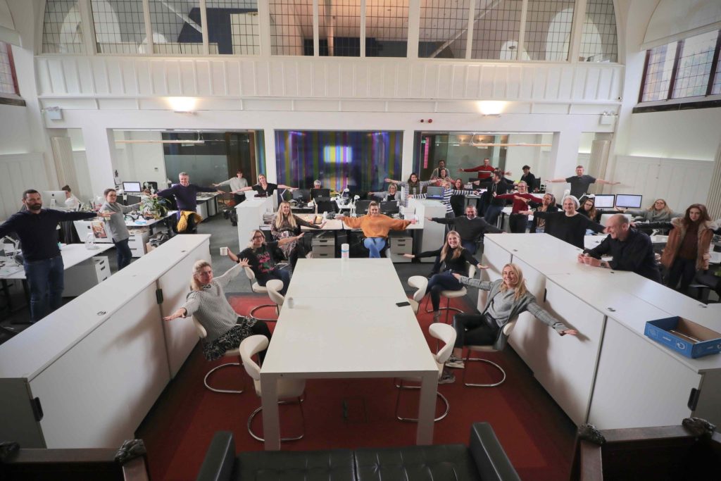 Office with group of people all standing or sitting at their desks looking to camera