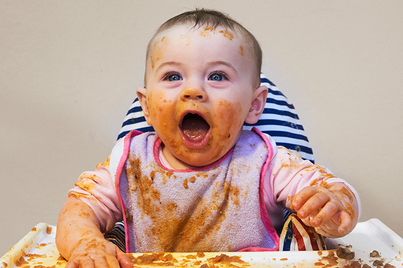 Baby eating, in high chair, with food all over it's face 