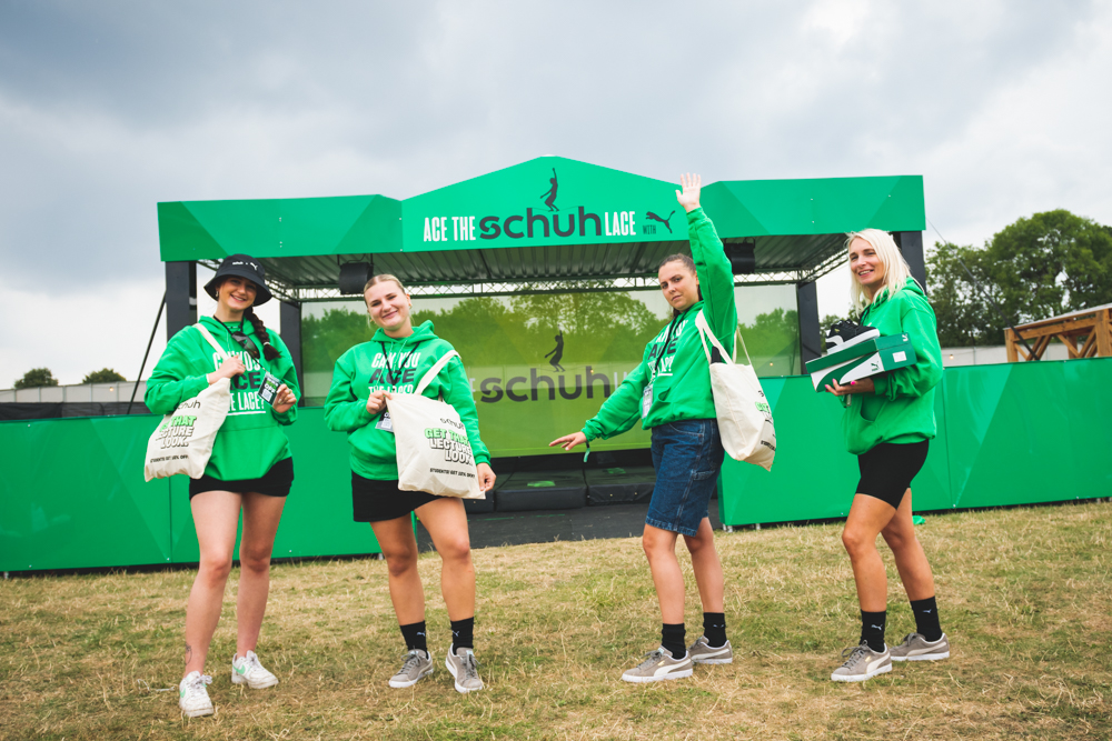 Four girls standning in field, in front of baner stand wearing green hoodies with logo and carrying branded logo bags