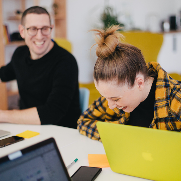 Man and woman sat at a desk in front of laptops, smiling and laughing