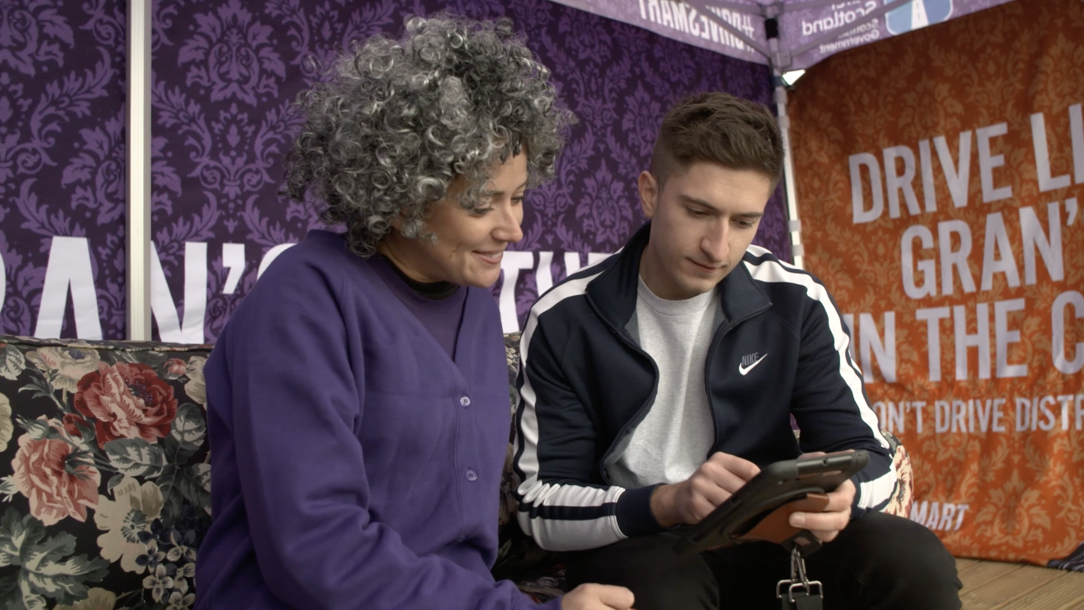 Woman dressed as old lady on sofa speaking to young man holding a tablet