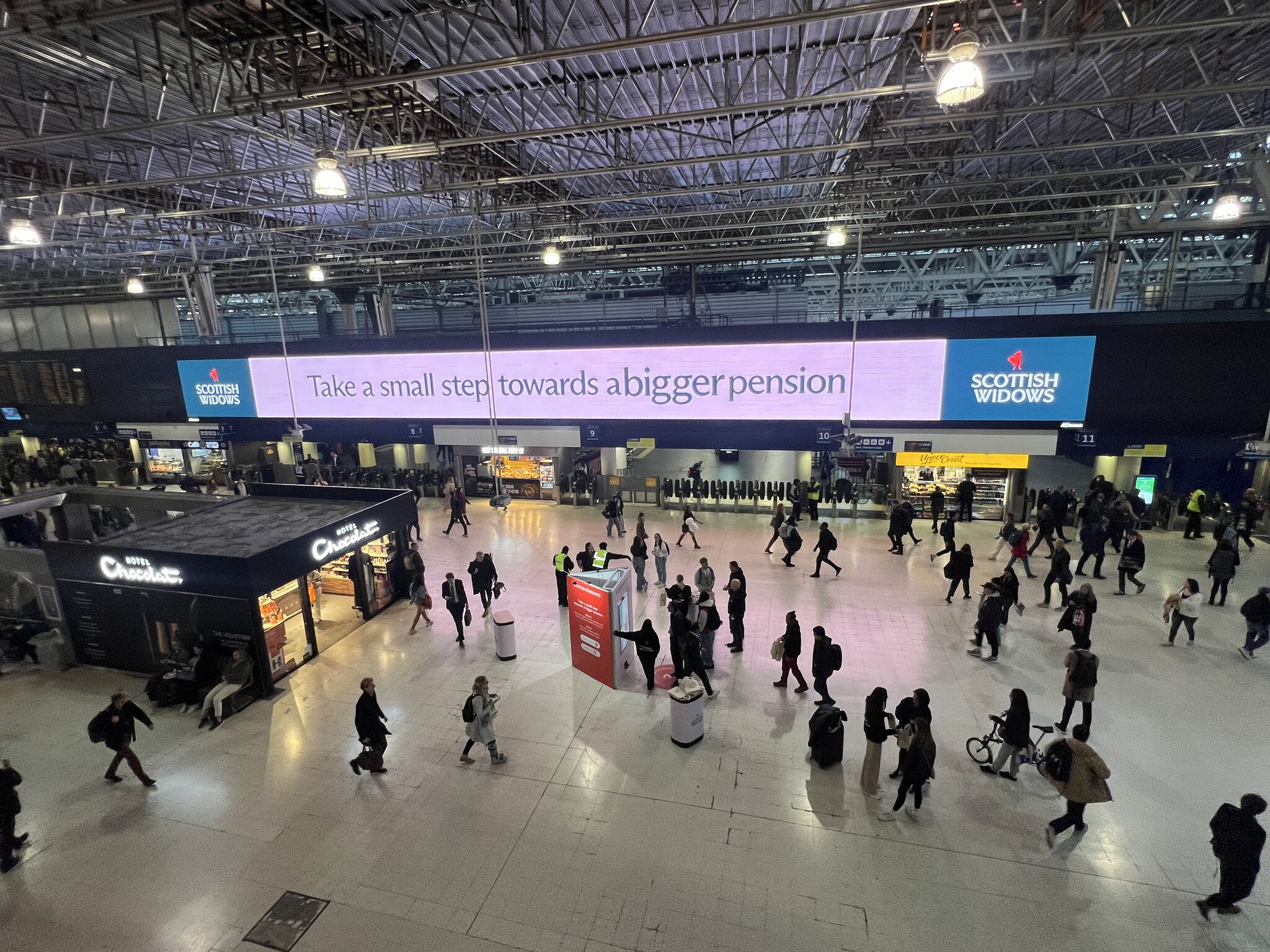 wide shot of the train station interior many people walking to and fro on platform and digital display standing in the middle being used