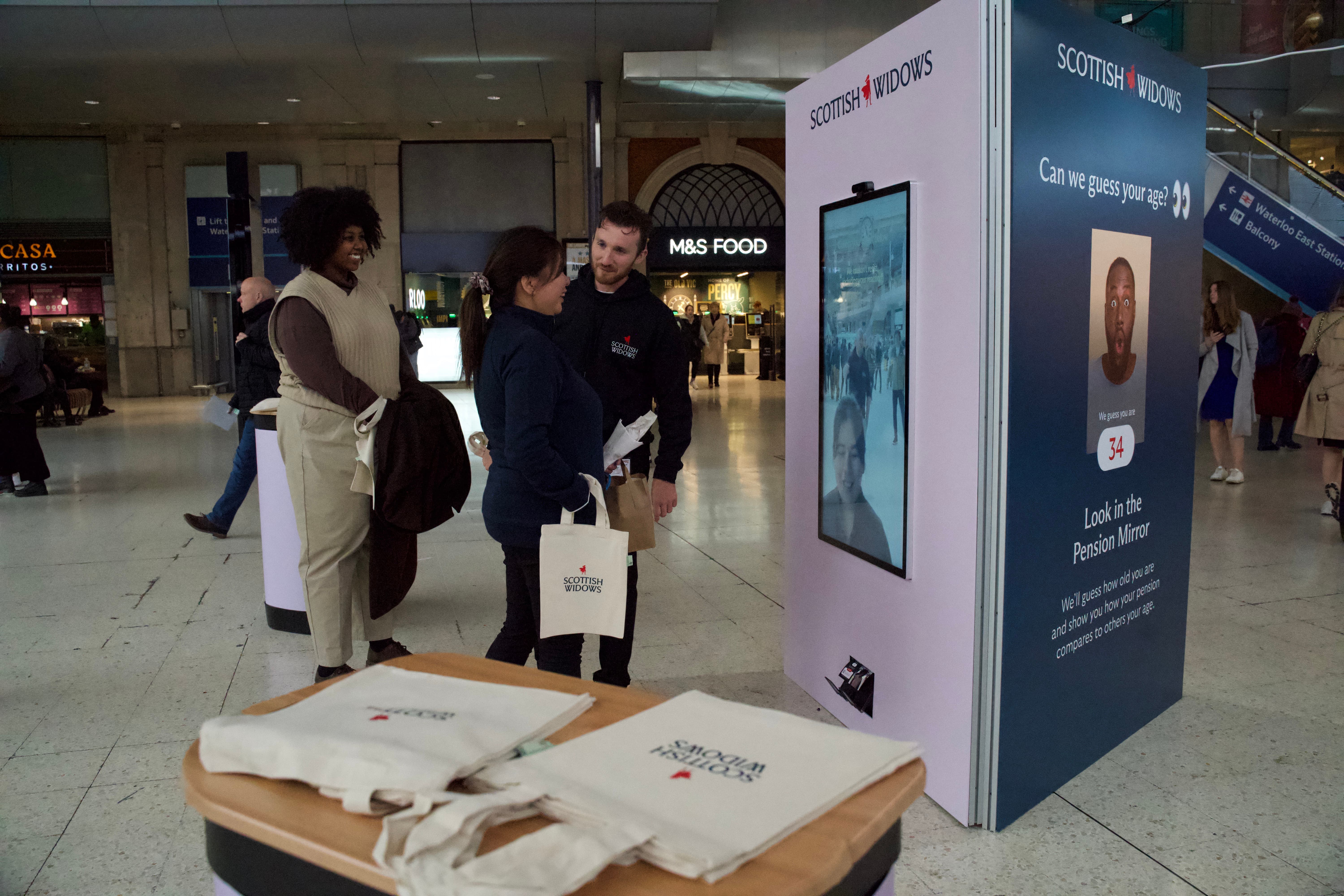 Man showing women how to use a digital display unit at train station