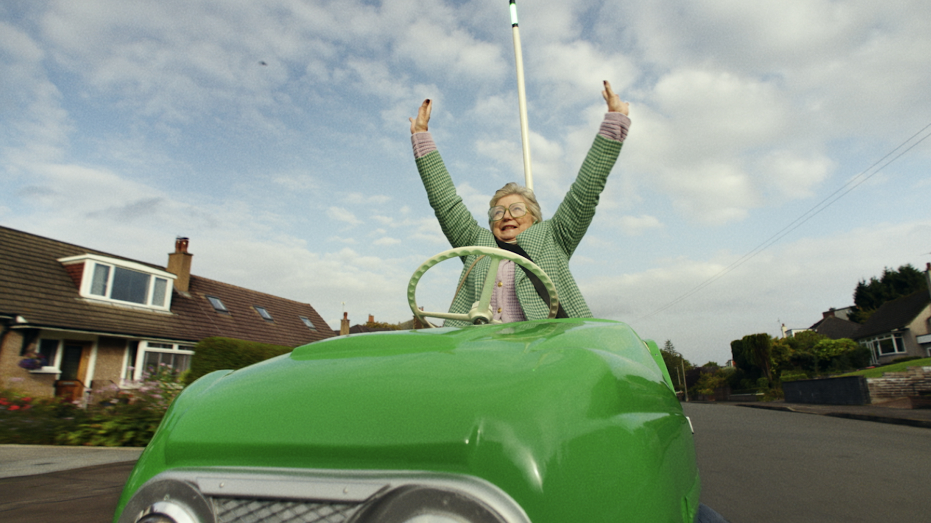 Old woman in a green bumper car driving on the street with her arms in the air